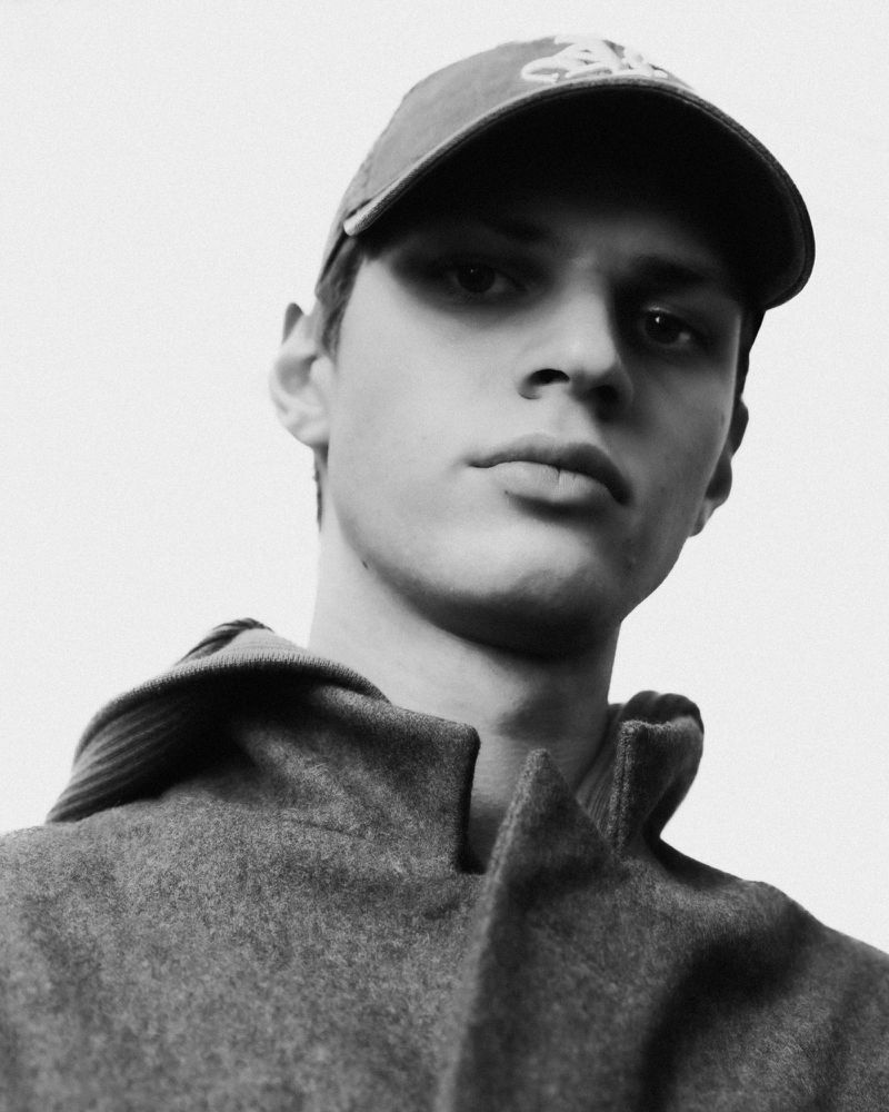 Monochrome portrait of a young man wearing a baseball cap and a heavy coat against a plain background.