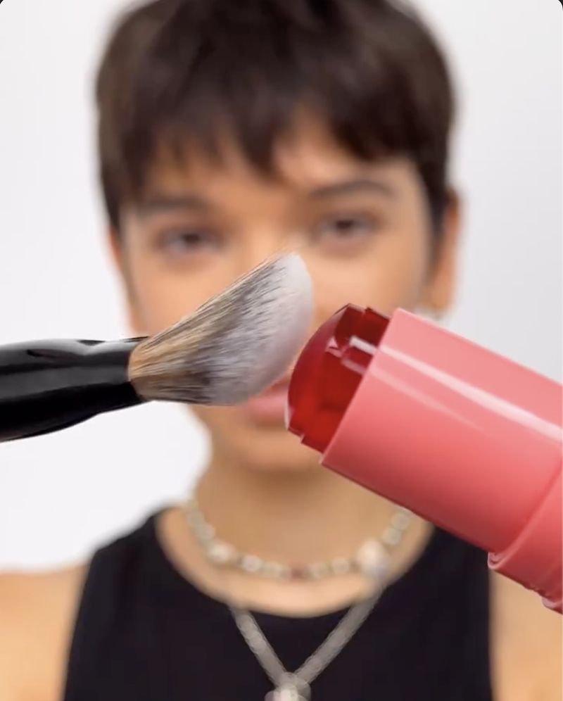 Close-up of adult with short hair applying a red-tinted balm with a cosmetic brush in a bright studio setting. Beauty concept.