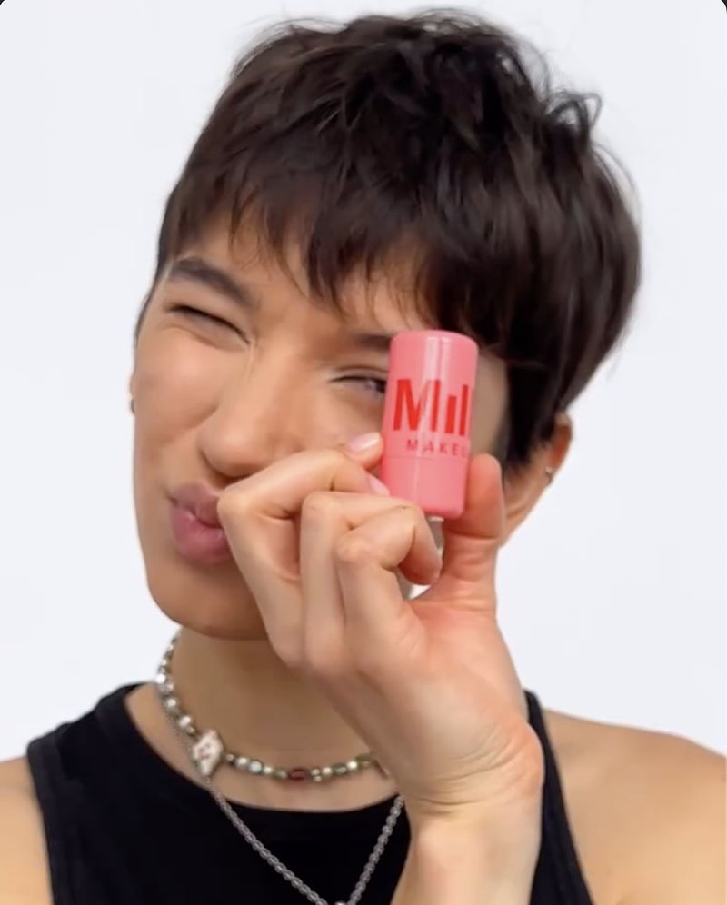 Woman with short brown hair is holding a Milk Makeup tube in front of a white backdrop, showcasing beauty and cosmetics.