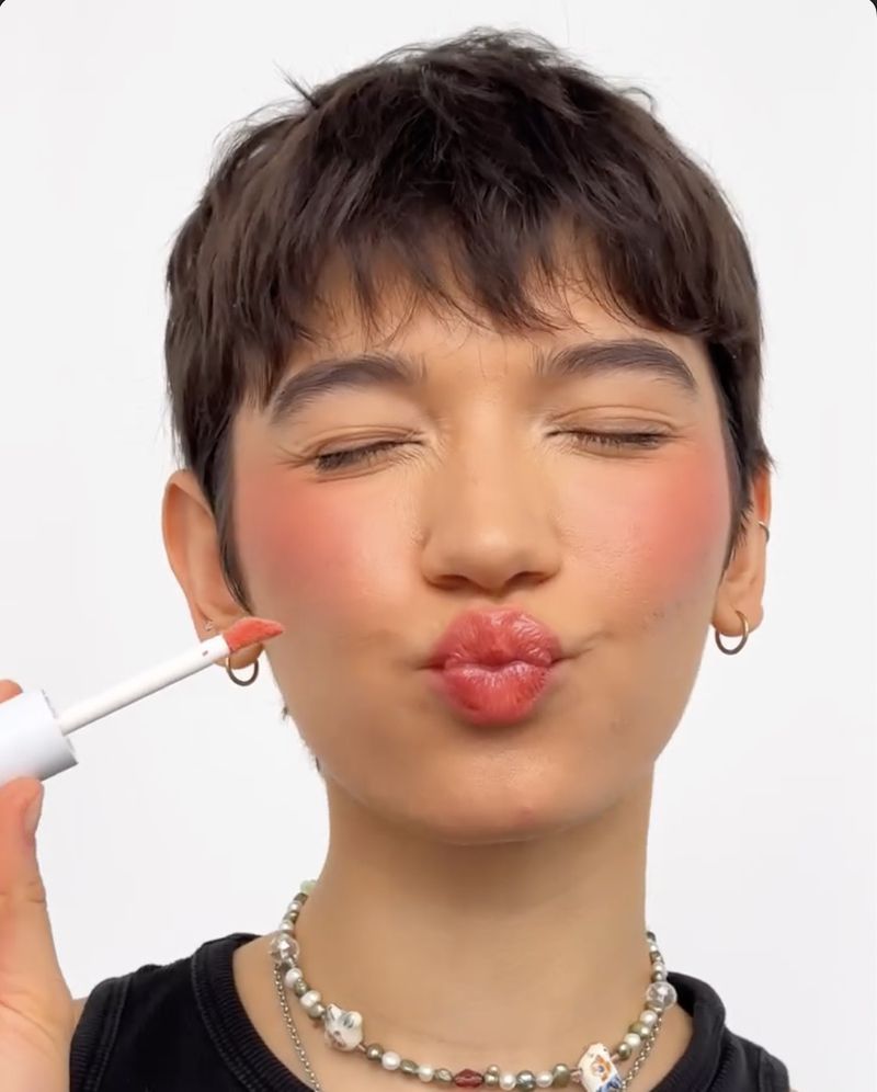 Close-up of a young adult woman applying lip gloss, with focus on her pouting lips and coral-toned blush, against a clean white backdrop.