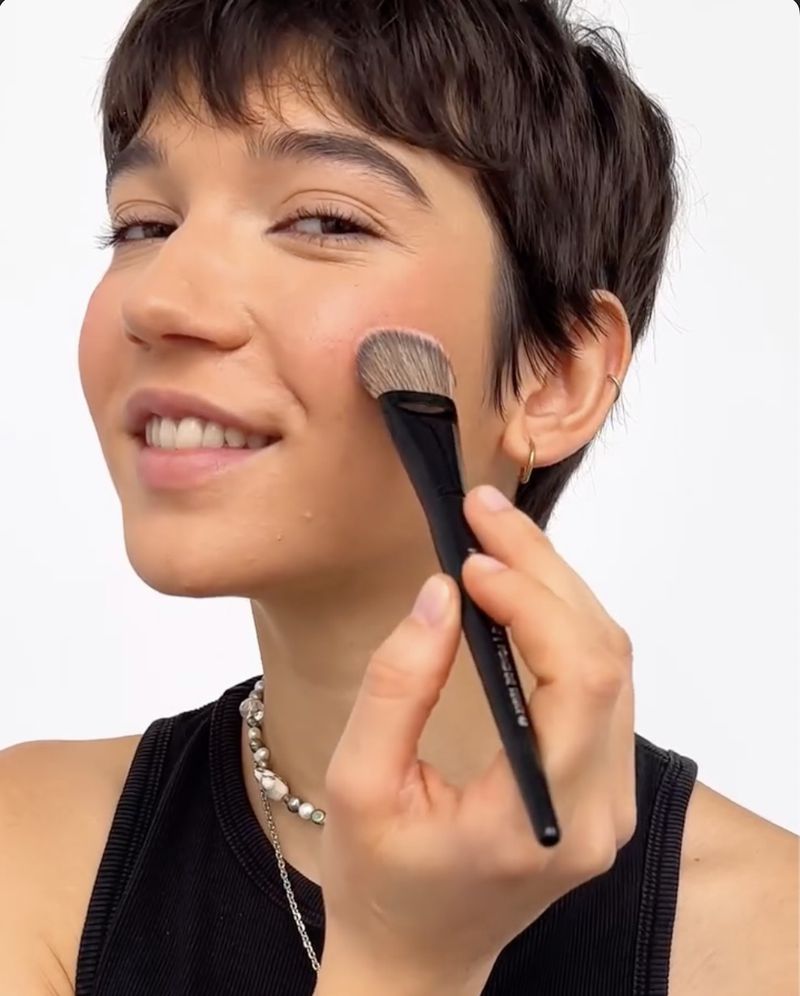A smiling woman applies blush with a cosmetic brush in a close-up studio shot. The focus is on her face and the makeup application.