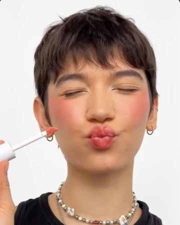 Close-up of a person with short brown hair pouting lips, holding a lip gloss applicator near the cheek.
