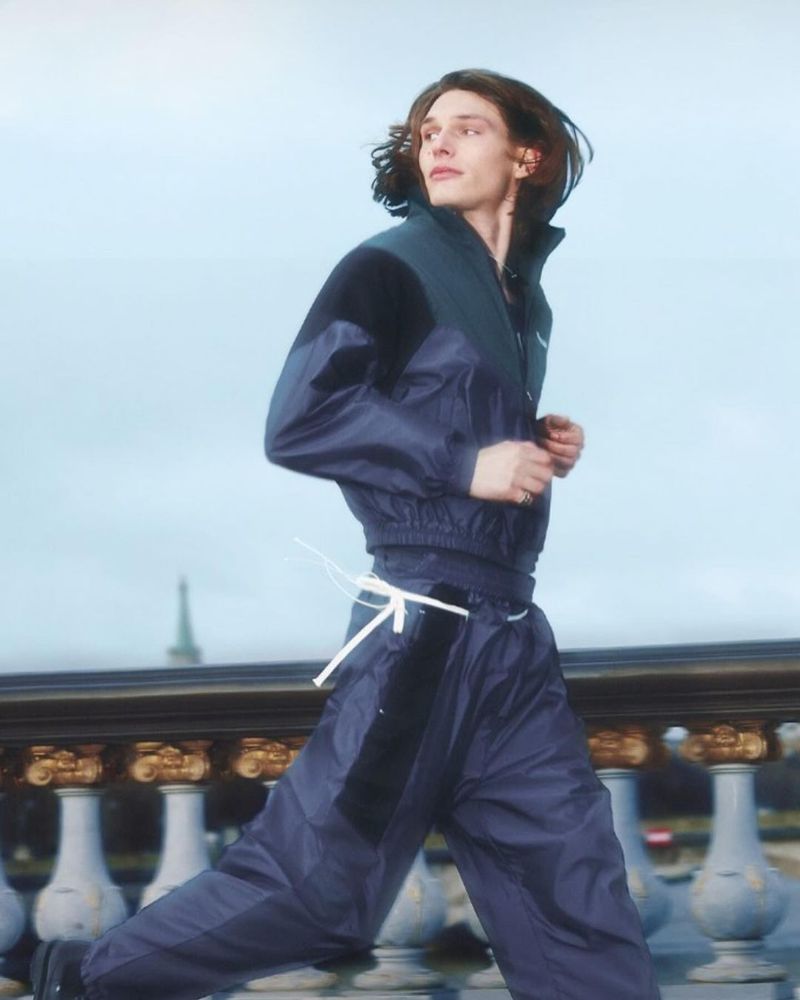 Male fashion model runs in blue nylon tracksuit near ornate stone balustrade under sky. Editorial fashion, lifestyle, outdoor, Parisian background.