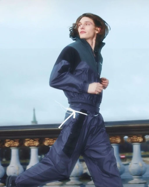 Male fashion model runs in blue nylon tracksuit near ornate stone balustrade under sky. Editorial fashion, lifestyle, outdoor, Parisian background.