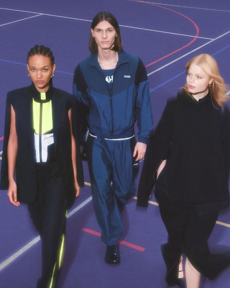 Three fashion models pose indoors on a blue court wearing athleisure outfits in black and blue. The models face the camera.