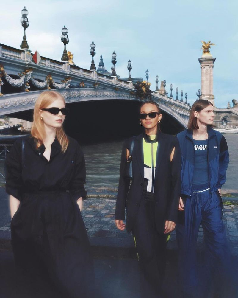 Three women in fashionable outfits pose near the Pont Alexandre III bridge in Paris, captured on a cloudy day.