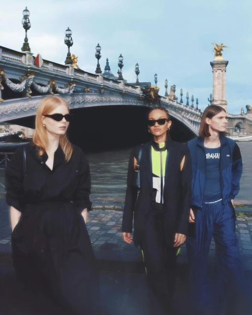 Three women in fashionable outfits pose near the Pont Alexandre III bridge in Paris, captured on a cloudy day.