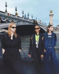 Three models walking along a riverside in Paris with a historic stone bridge in the background.