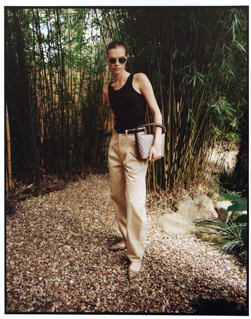 A slender woman with sunglasses poses outdoors in a black top, khaki trousers, and heeled shoes, holding a patterned handbag near bamboo.