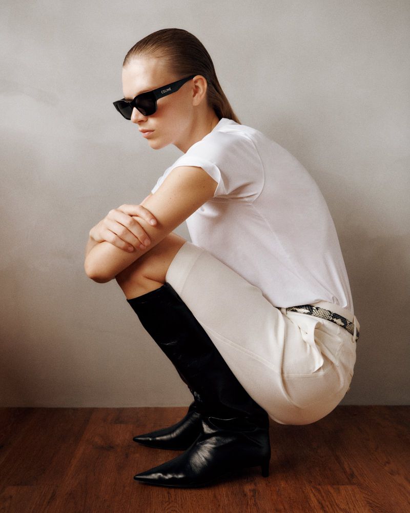 A woman in a white T-shirt, white shorts, and black knee-high boots poses in front of a muted grey wall on a wood floor.
