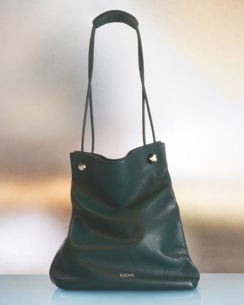 Dark green Loewe shoulder bag featuring a top handle and a tonal logo on a light blue surface, captured in a studio setting.