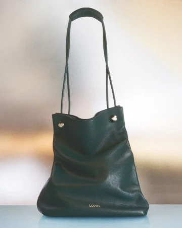 Dark green Loewe shoulder bag featuring a top handle and a tonal logo on a light blue surface, captured in a studio setting.