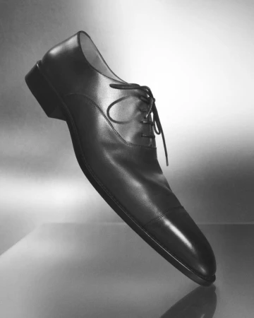 Black leather oxford dress shoe on a reflective surface against a grey gradient background in a studio setting, men's fashion.