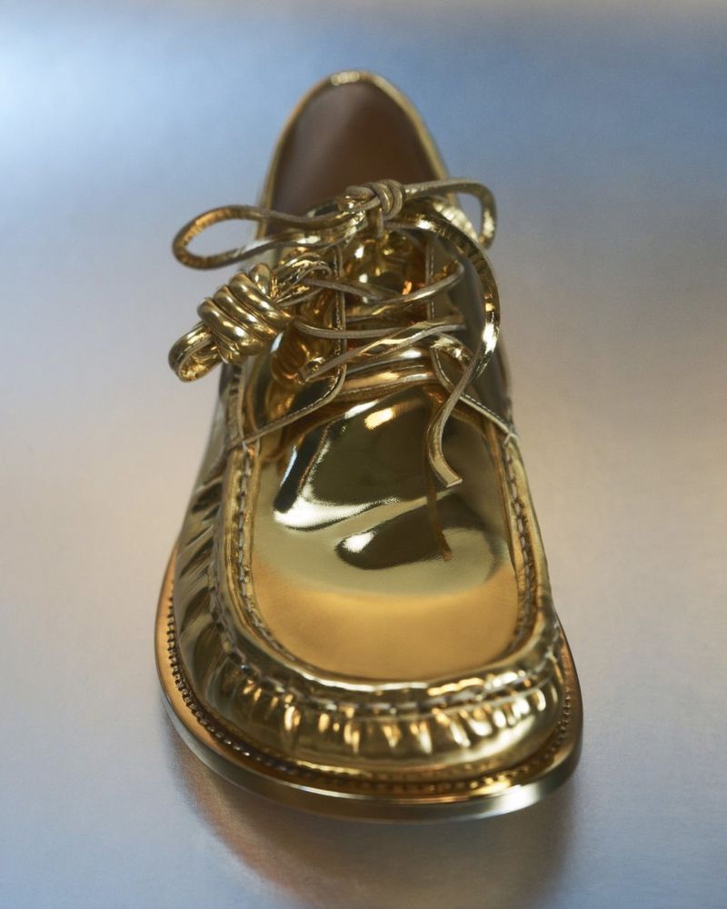 A glossy gold-colored shoe with laces is displayed up close, exhibiting high shine and reflective surfaces set against a gradient background.