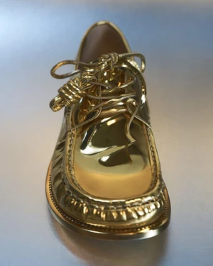 Close-up product shot of a metallic gold leather loafer shoe with tied laces resting on a grey seamless background.
