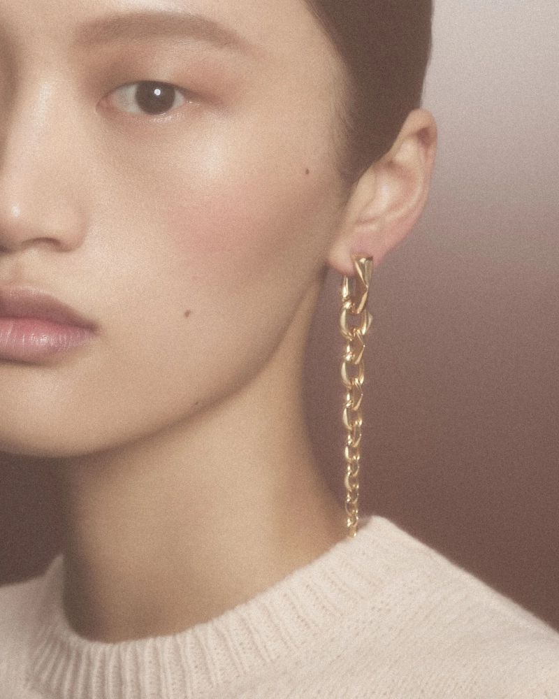 Close-up portrait of a model wearing a long gold chain earring and a white textured knit sweater against a warm neutral background.