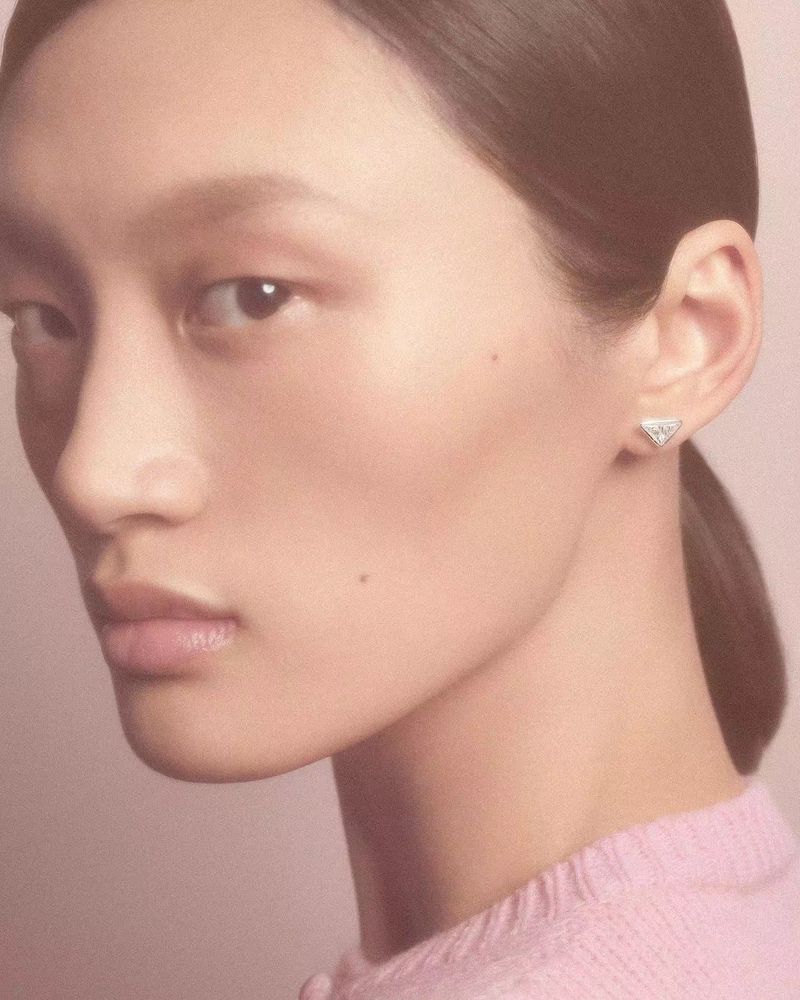Portrait of an adult female with light skin, wearing pink, and featuring a small, triangular silver earring, in a soft, evenly lit studio setting.