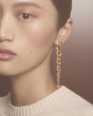 Close-up portrait of a model wearing a long gold chain earring and a white textured knit sweater against a warm neutral background.