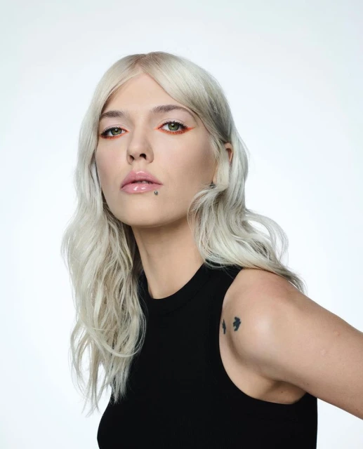 Close-up portrait of a woman with blonde hair, orange eyeliner, and a black top against a white background. Beauty shot.