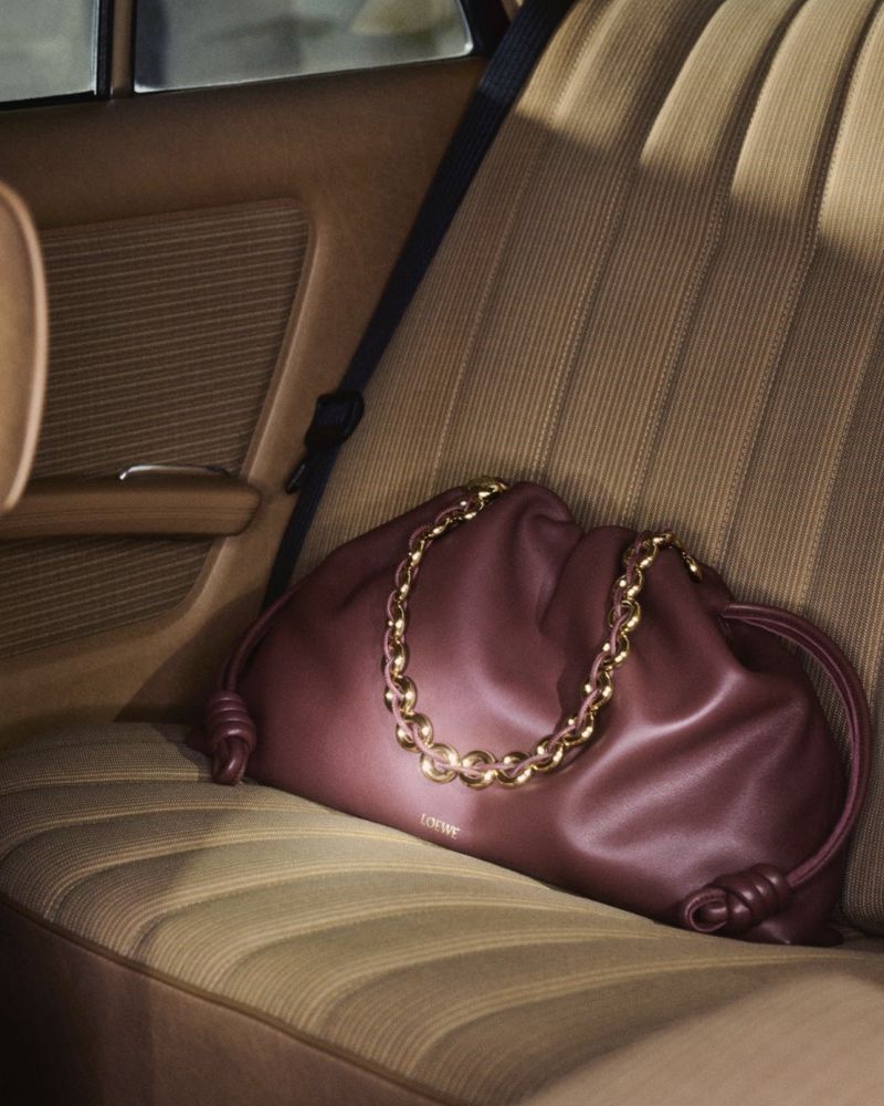 Burgundy leather handbag with gold chain detail on a tan striped car seat with a black seatbelt, luxury fashion accessory.