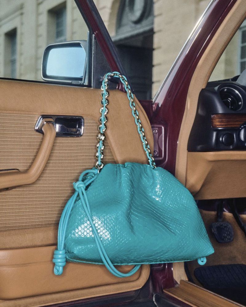 A turquoise purse with a gold chain hangs on the open door of a luxury car, showcasing fashion and automotive elements with an urban backdrop.