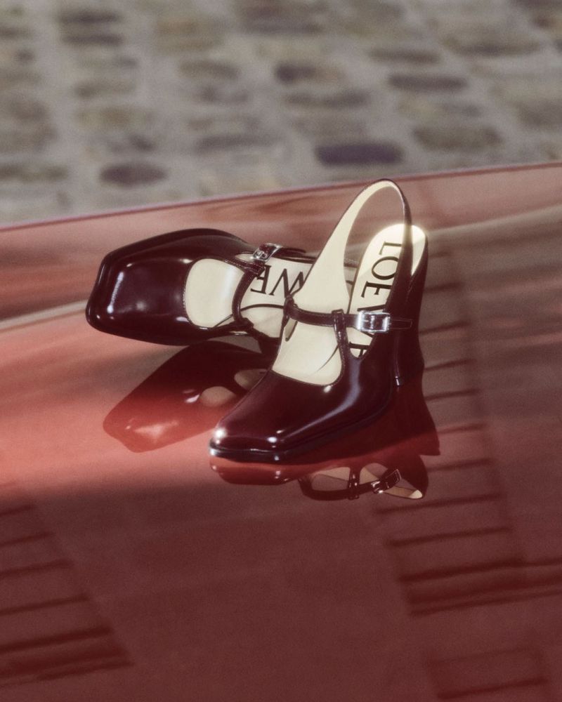 Pair of patent leather Mary Jane shoes by Loewe, one angled on its side displaying the logo, on a reflective red surface.