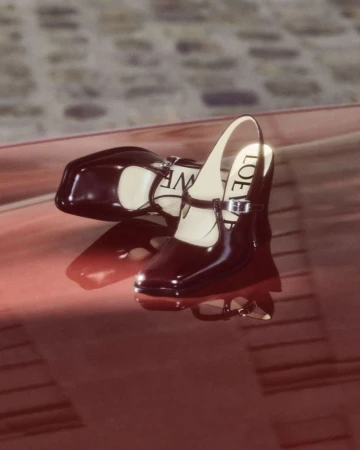 Pair of patent leather Mary Jane shoes by Loewe, one angled on its side displaying the logo, on a reflective red surface.