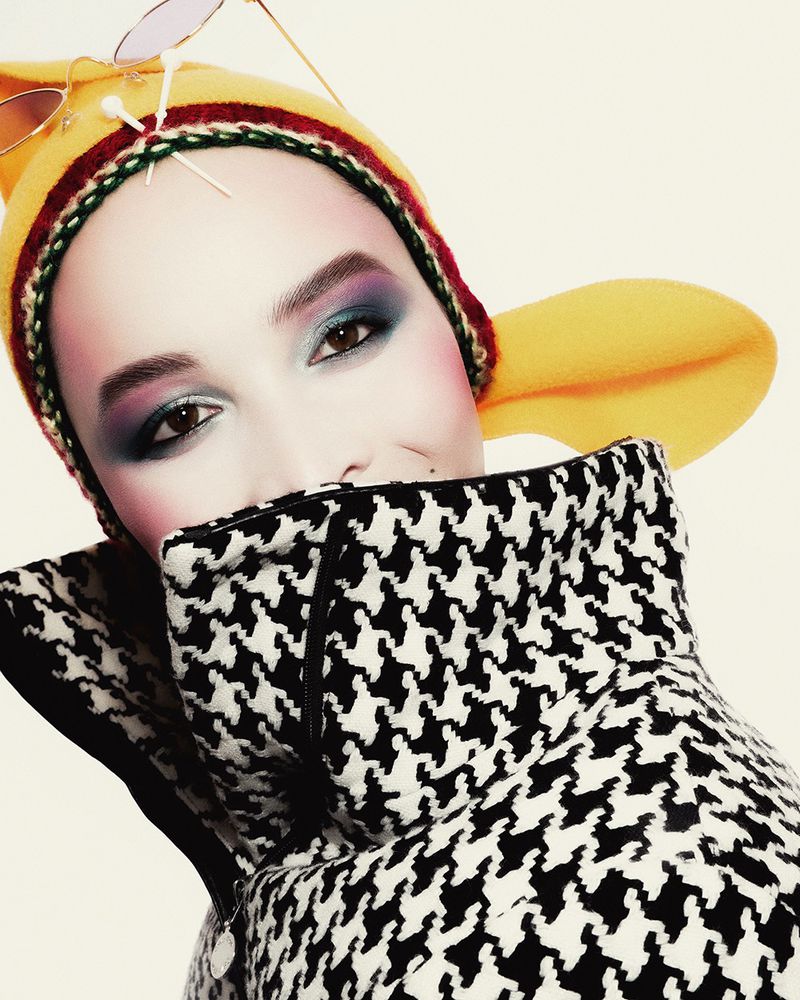 Close-up of a woman wearing a yellow hat and black and white patterned jacket, striking pose with bold eyeshadow and pink blush.