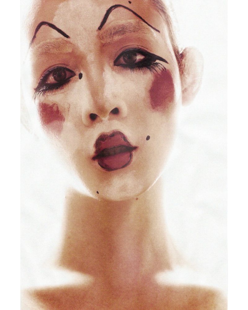Close-up portrait of a person with dramatic clown-like makeup, featuring heavy eyeliner, arched eyebrows, and accentuated red lips and cheek color.