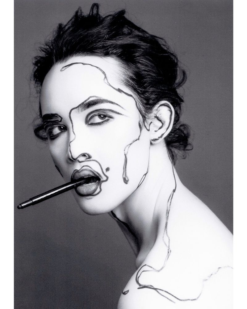 Monochrome portrait of a woman with graphic black lines drawn on her face and neck, holding a black pen in her mouth.