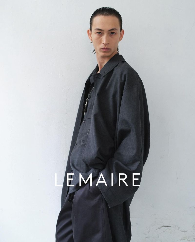 Adult male poses in loose dark grey shirt and pants against a light grey wall. The fashion brand 'LEMAIRE' is overlaid on the image.