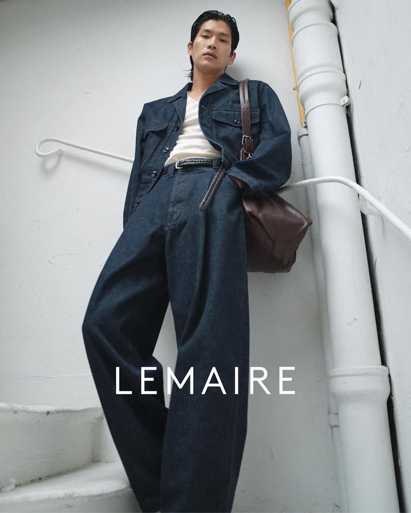 An Asian male model showcases a denim jacket and baggy jeans with a brown leather bag in a fashion-forward setting against a neutral wall.