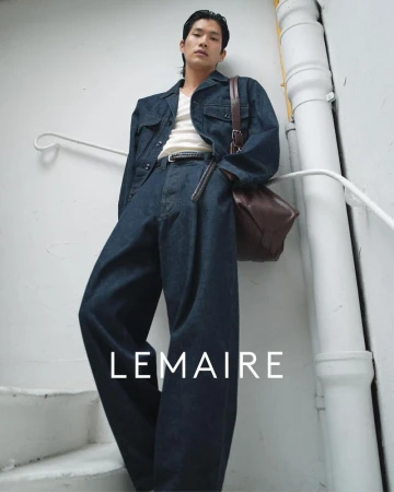 An Asian male model showcases a denim jacket and baggy jeans with a brown leather bag in a fashion-forward setting against a neutral wall.