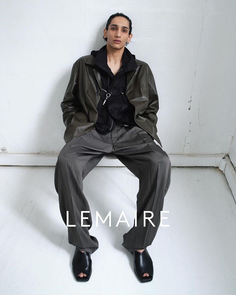 Male model in an olive jacket and gray pants sits in a studio against a white wall, showcasing a relaxed and fashionable pose with brand logo.