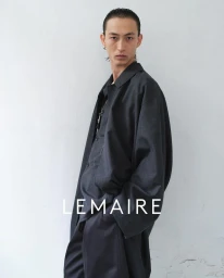 Model wearing dark oversized coat against a white wall with Lemaire text overlay