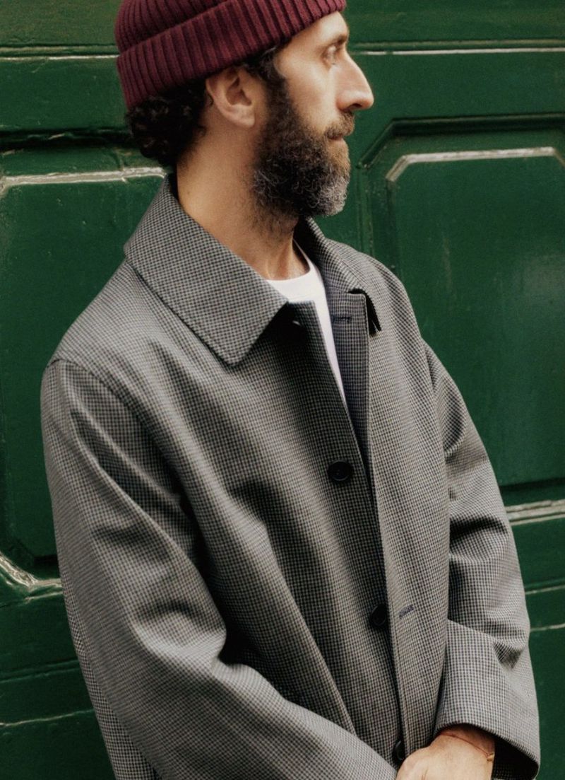 Profile shot of a bearded adult male wearing a grey coat and maroon beanie, standing against a green wall outdoors, fashion editorial.