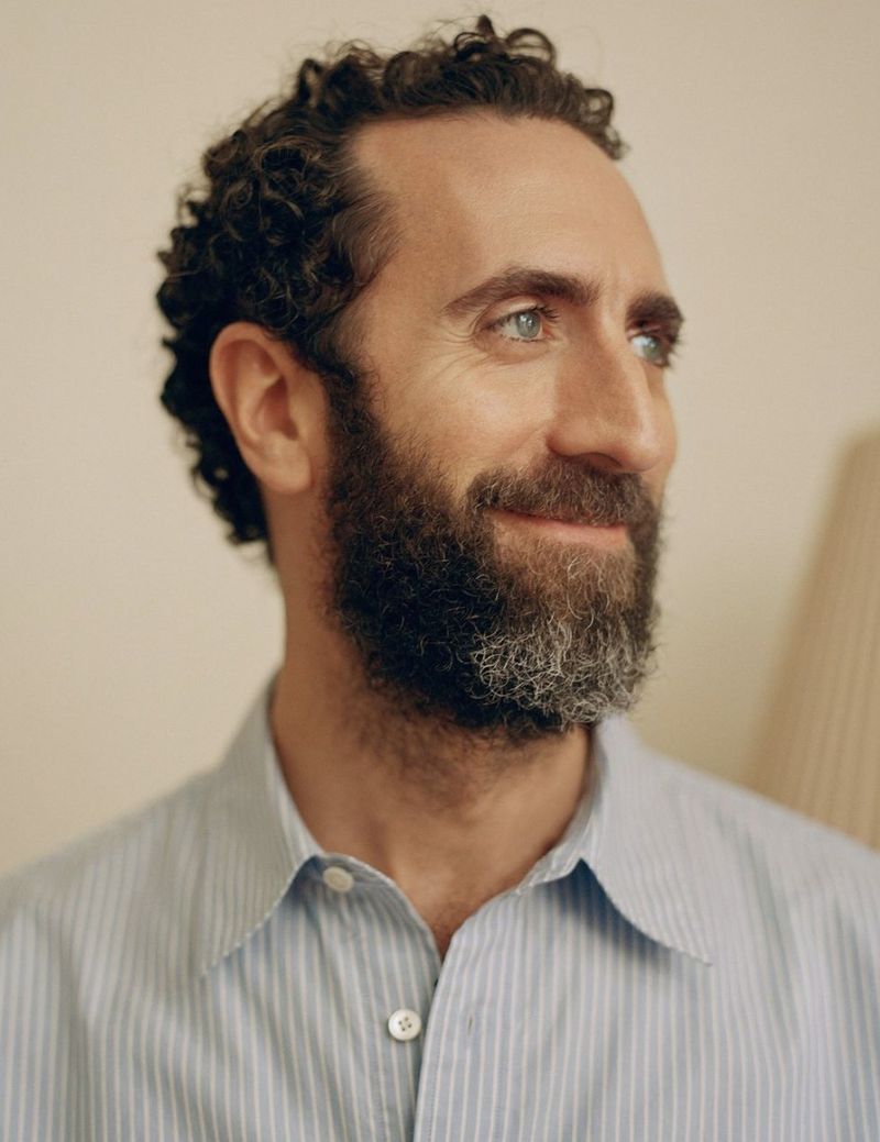 Portrait of a man with short curly dark hair and a beard wearing a light blue striped collared shirt against a neutral background.