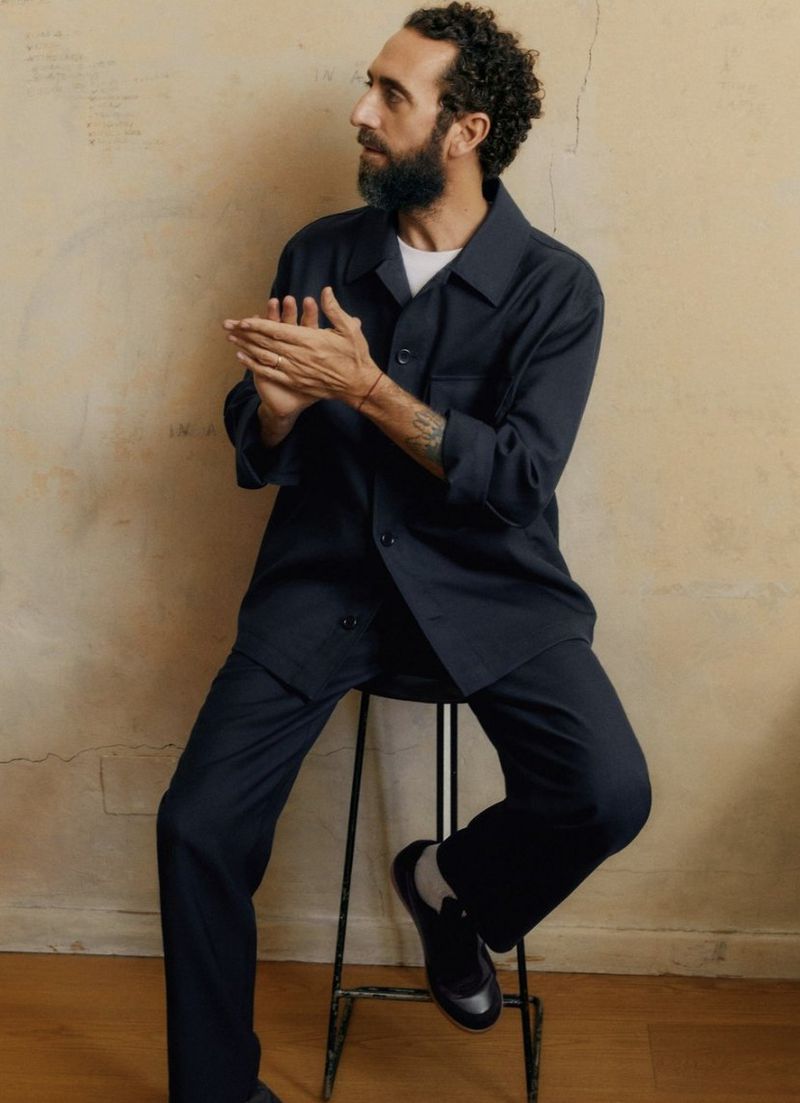 Man with beard seated on a metal stool wearing a dark navy jacket and trousers against a textured wall