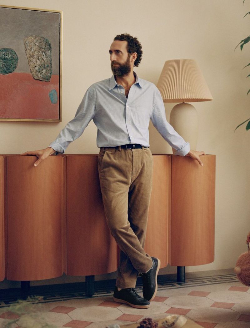 Bearded man in a blue shirt and brown pants stands near a wooden cabinet with a lamp and painting in a well-lit room.