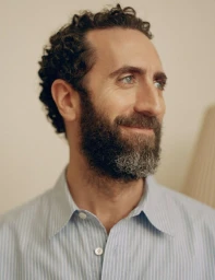Portrait of a man with short curly dark hair and a beard wearing a light blue striped collared shirt against a neutral background.