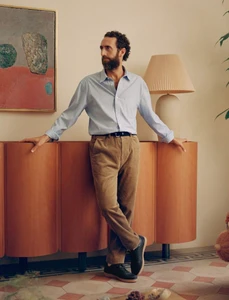 Bearded man in a light blue shirt and brown trousers leaning against a wooden cabinet in a room