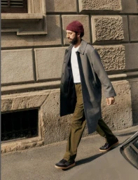 Man wearing a grey coat, white t-shirt, and beanie walking past a rustic stone building facade