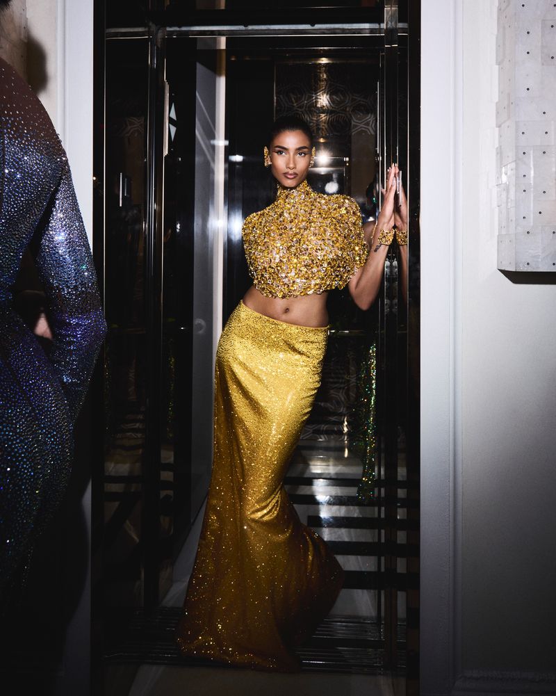 Glamorous woman in a gold sequin gown poses in a mirrored elevator, exuding elegance and style with subtle reflections and soft lighting.