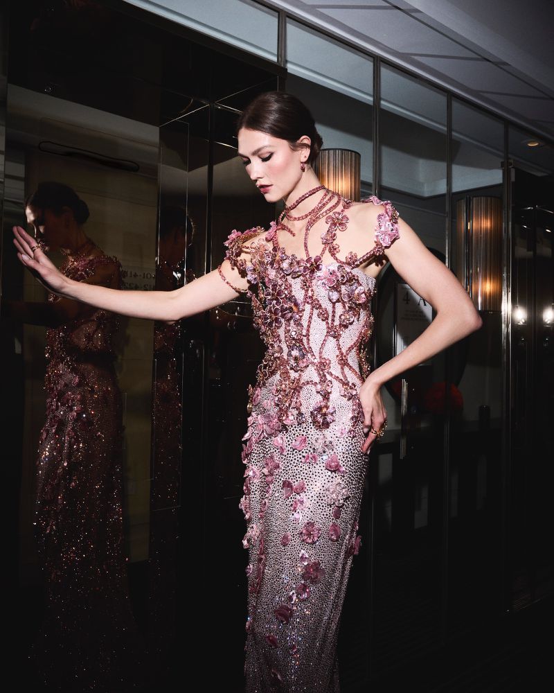 Elegant woman in a pink floral gown poses against a mirrored wall, showcasing intricate details of the dress and her sophisticated jewelry.