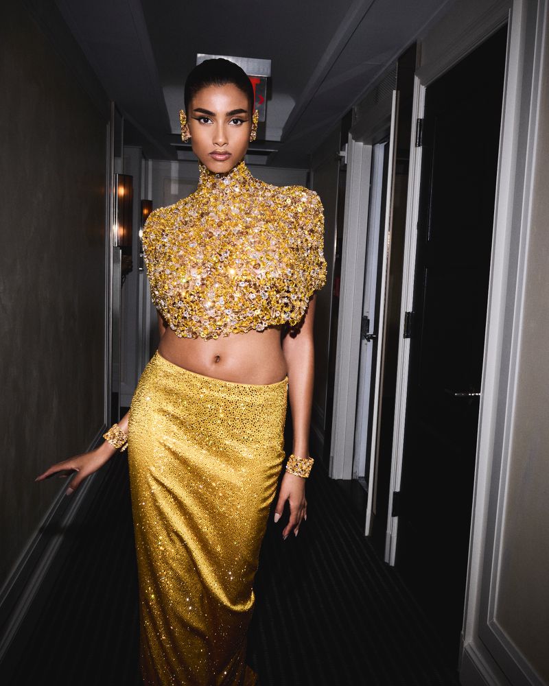 Woman in a gold, beaded crop top and skirt stands in a hallway. She poses with her arms down and wears gold bracelets.
