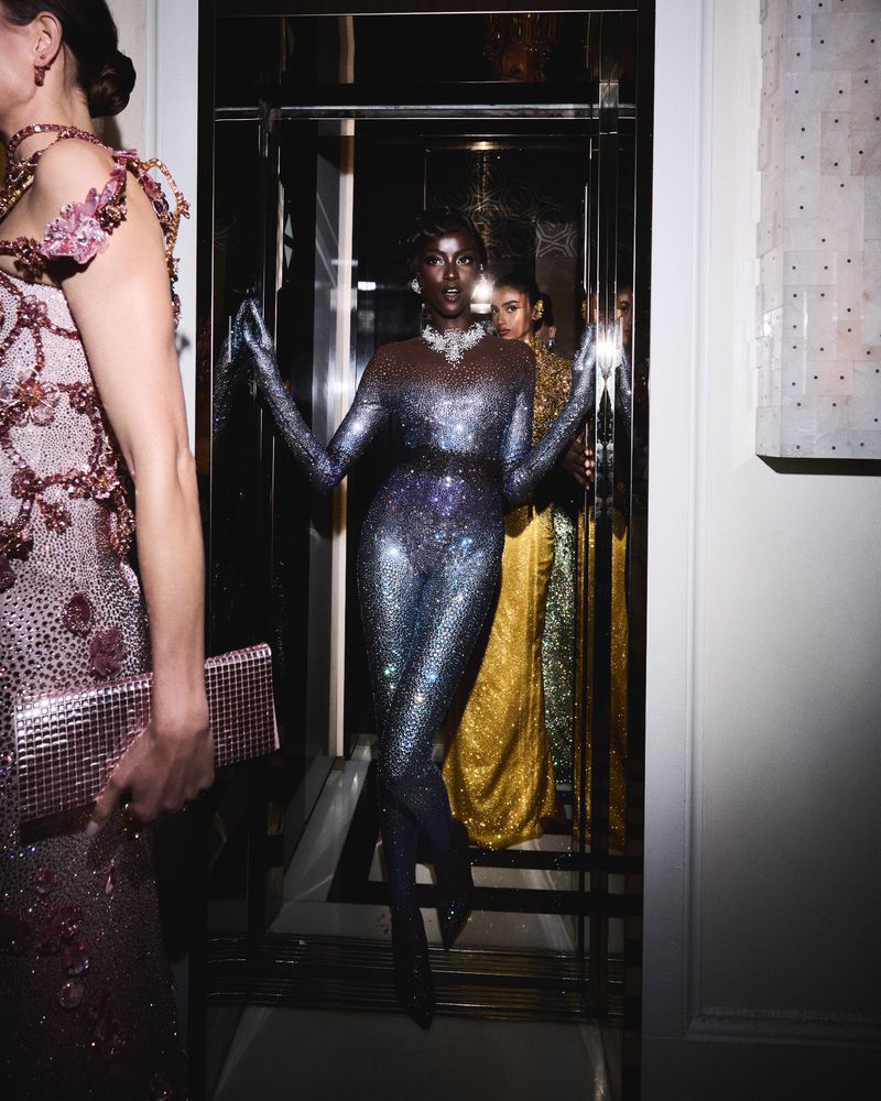 Runway models wearing sequined gowns walk through a doorway. One model wears a sparkling jumpsuit, necklace, and heels.