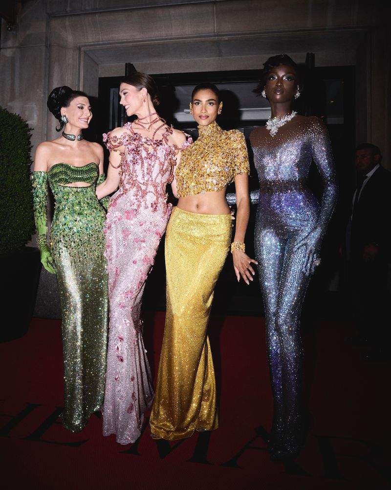 Four fashion models in shimmering formal gowns on a red carpet. The women are wearing glamorous jewelry. Outdoor setting, dark lighting.