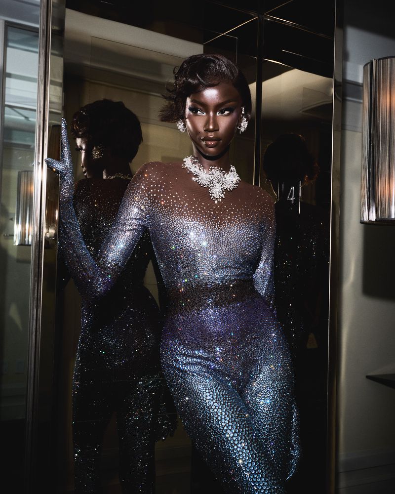 Elegant Black woman in shimmering jumpsuit and jewelry strikes a pose in an indoor setting near a reflective surface and decorative lamp.