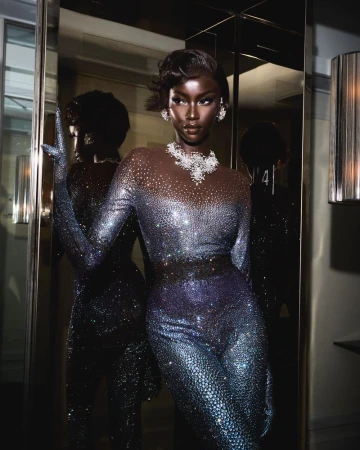 Elegant Black woman in shimmering jumpsuit and jewelry strikes a pose in an indoor setting near a reflective surface and decorative lamp.