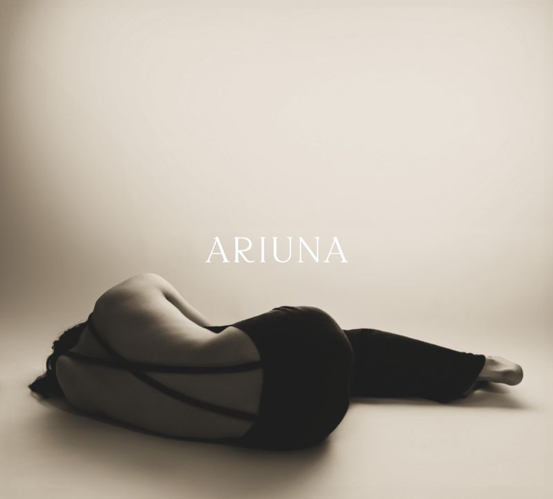 An adult female in dark clothing reclines against a light backdrop, with her face obscured and body curled, bathed in studio lighting.